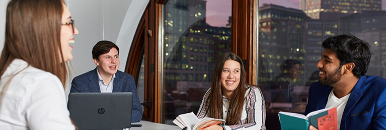 A group of students with a laptop at a table with a window looking out onto highrise buildings