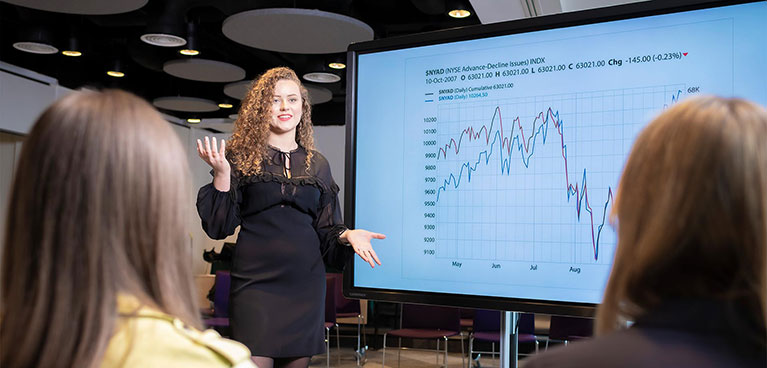 Female presenting beside a screen with graphs with 2 students looking on 