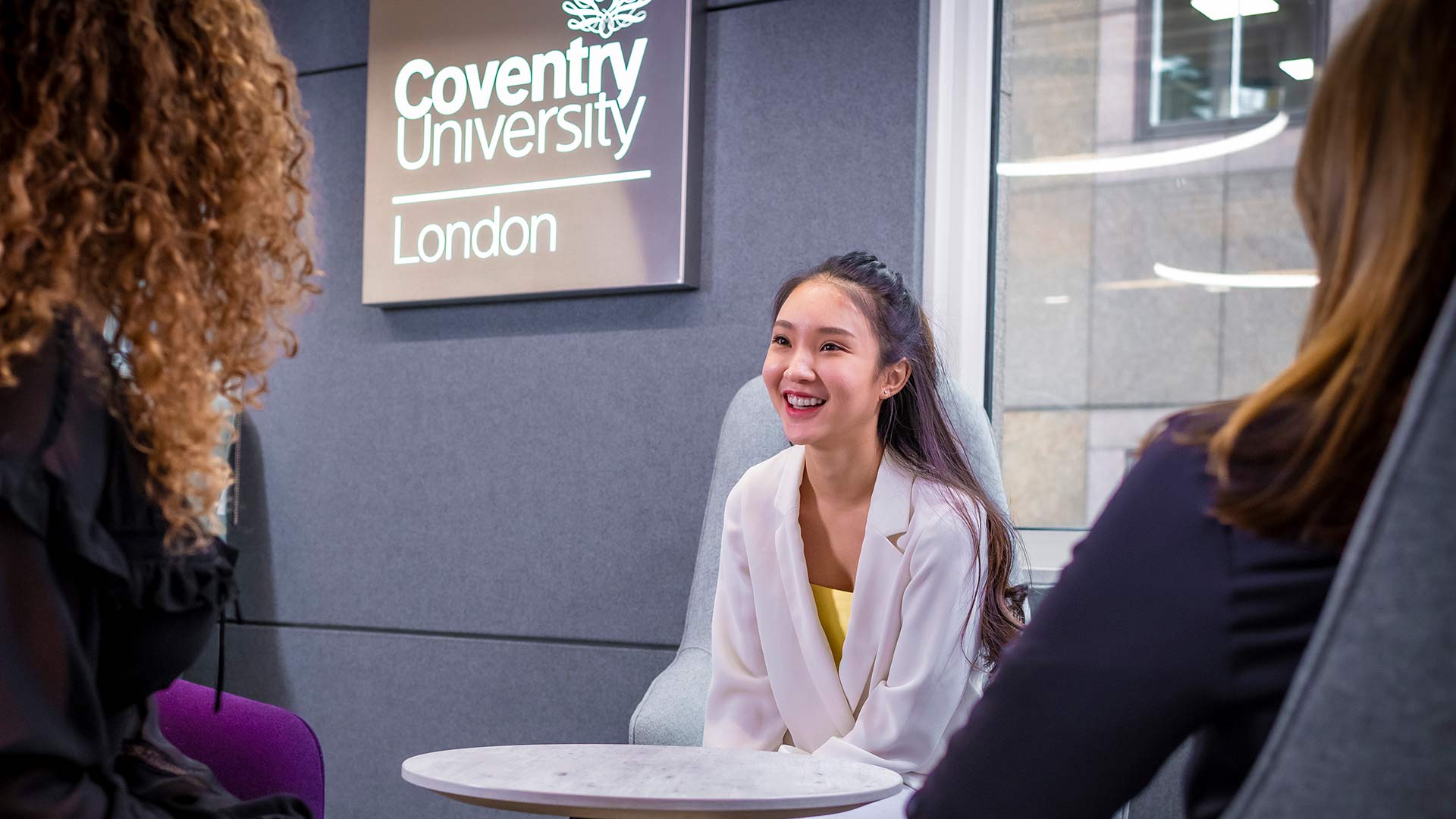 young female student chatting to two other students with the coventry university london sign on the wall in the background 
