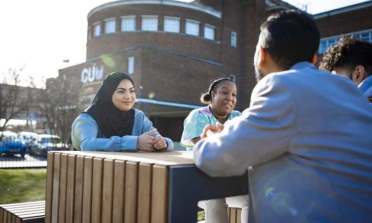 Students smiling and chatting while sat down at a bench outside a CU London building which can be seen in the background