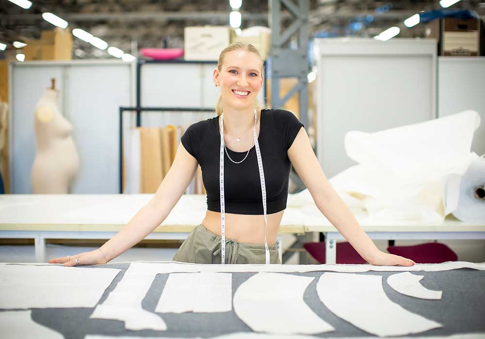 smiling female student with a measuring tape around her neck standing at a table with dressing making cut outs