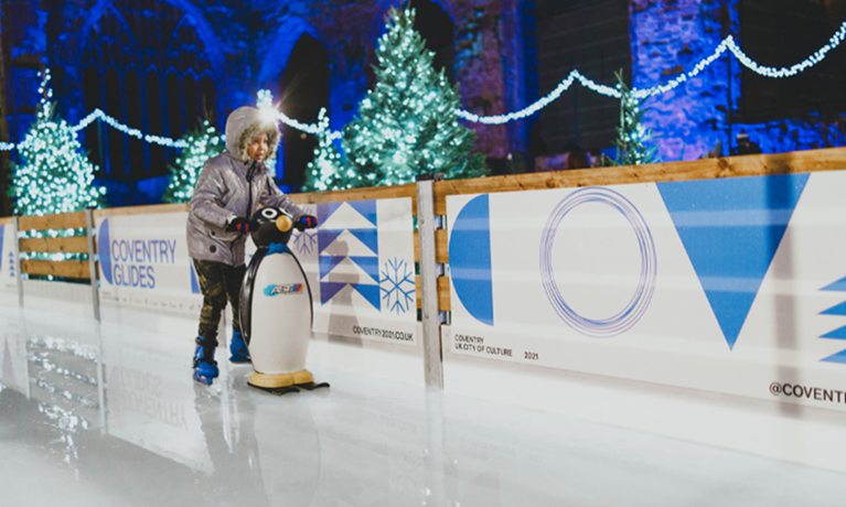 A child ice skating using a penguin styled stabiliser with Coventry Cathedral up lit with a blue light