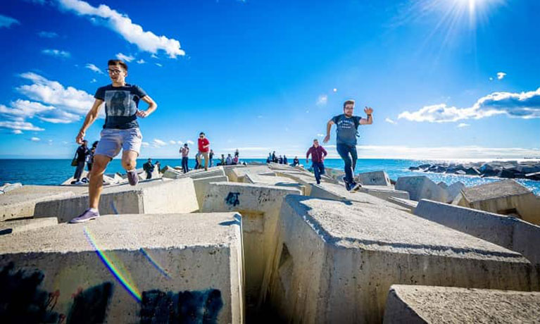 group of young students running on large boulders with blue sky and sea in the background