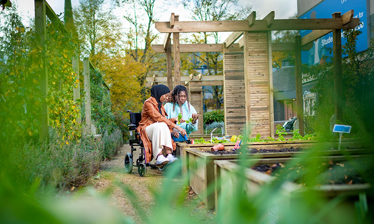 A woman in a wheelchair sits next to another woman amongst a herb garden as they hold one of the plants.