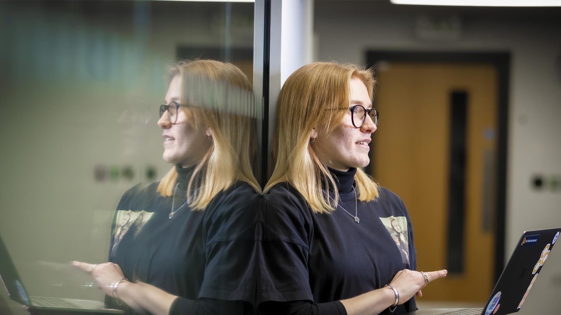 A cloud computing student with a laptop leaning against a glass wall that she is reflected in.