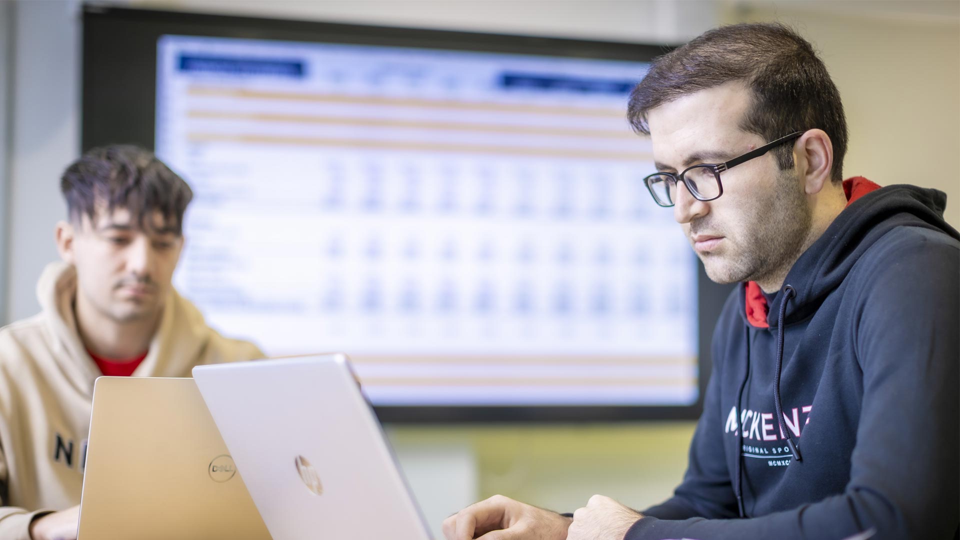 Two students working on laptops in front of a large wall-mounted screen.