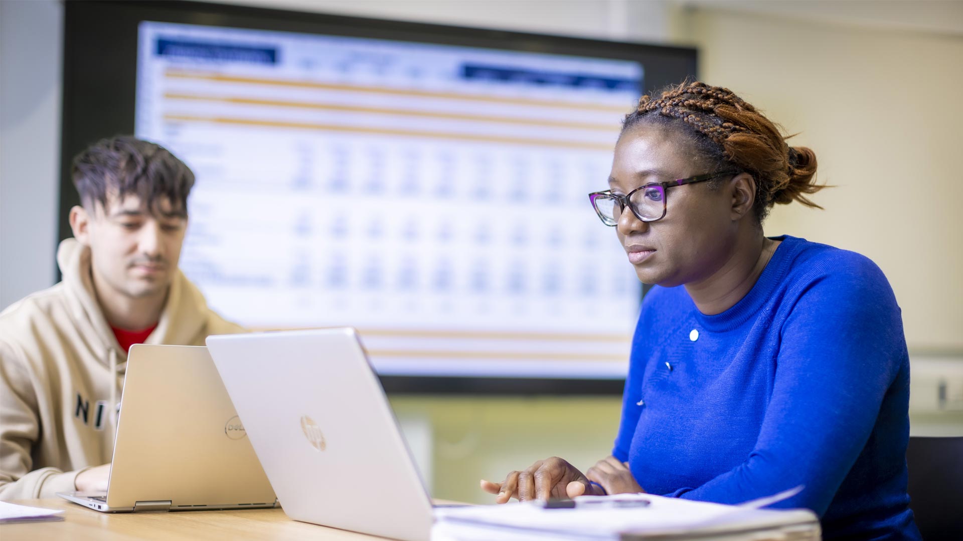 Two students working on laptops in front of a large wall-mounted screen.