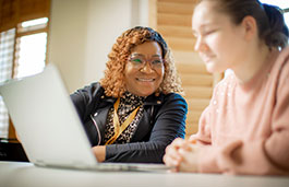 A smiling lecturer looks at a student who is concentrating on her laptop