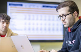 Two students working on laptops in front of a large wall-mounted screen.