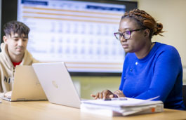 Two students working on laptops in front of a large wall-mounted screen.