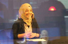 Smiling student stood in front of a TV screen wearing a yellow hijab