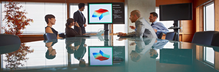 Four smartly-dressed people sitting at a table talking, while a fifth looks at information on a large screen