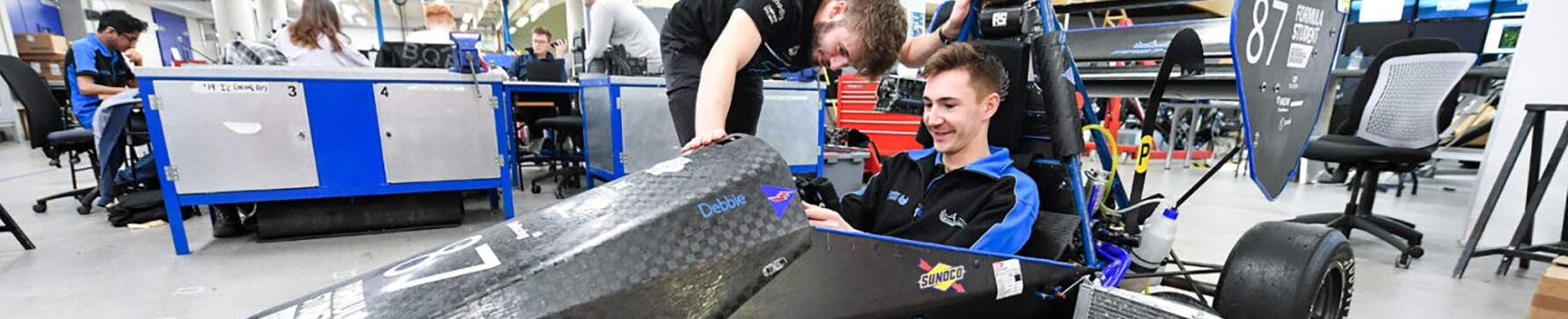Smiling student sitting in motorsport car with another student looking on