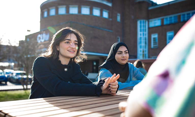 Two female students sat outside on a bench talking