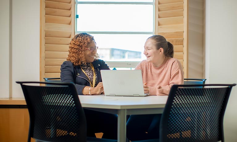 Two students sat at a desk talking in front of a laptop