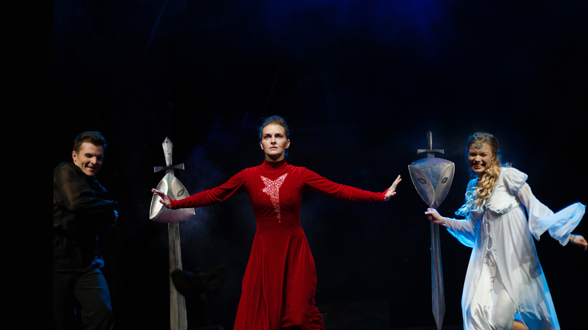 Three actors on stage, the centre one in a dramatic stance, the other two smiling on either side in the background