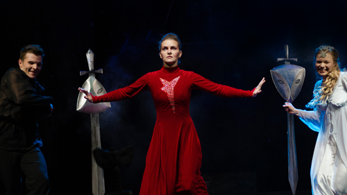 Three actors on stage, the centre one in a dramatic stance, the other two smiling on either side in the background