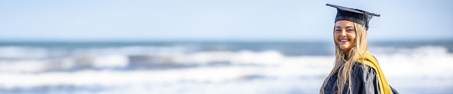 Female graduate in her graduation gown and cap stood in front of the sea