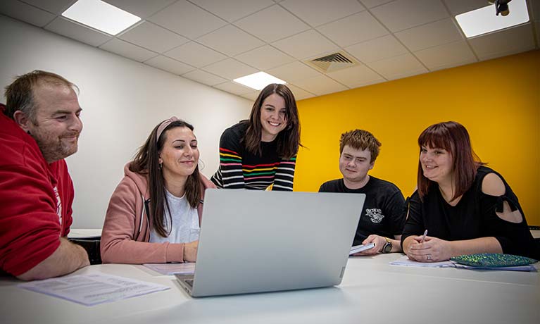 Group of students sat at a table with a lecturer standing next to them talking