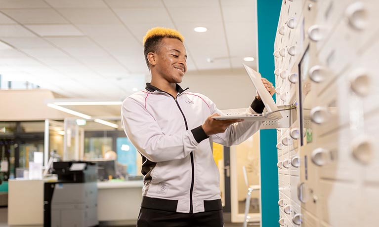 Male student stood next to laptop cupboard holding a laptop