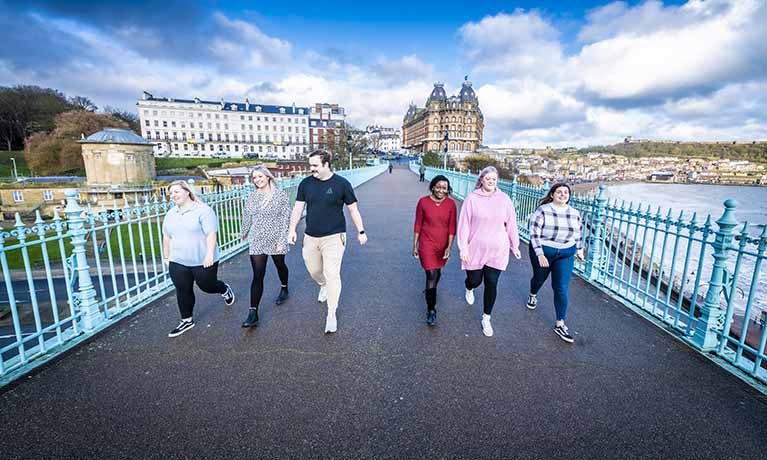 Group of students walking on the pier with the beach and Scarborough in the background.