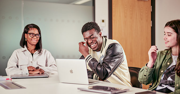 Three students sat at a desk with an open laptop laughing