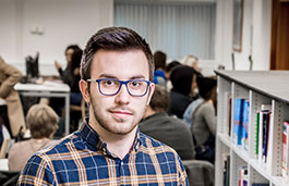 Man with glasses standing in a library next to a bookshelf