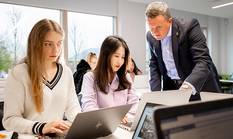 Student working on a laptop with a lecturer talking to a student behind