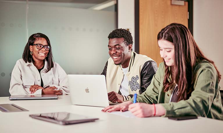 Three smiling students sat at a desk looking at a laptop 