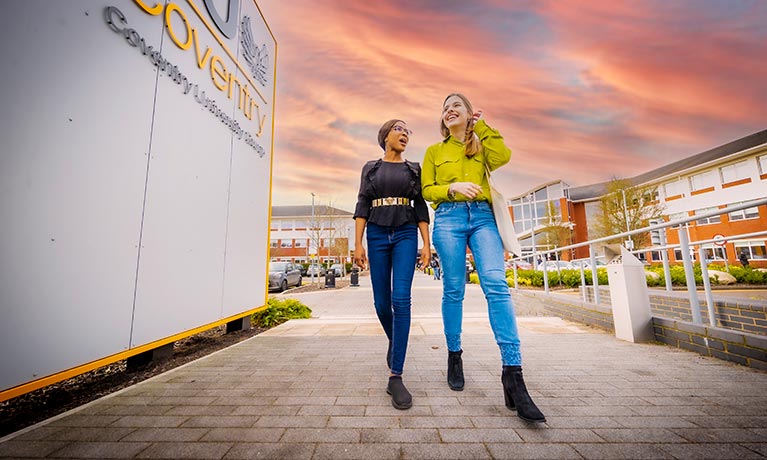 Two students walking past the CU Coventry building with a pink orange sky behind them