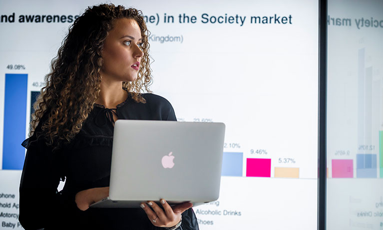 Woman presenting holding a laptop in front of a whiteboard of graphics