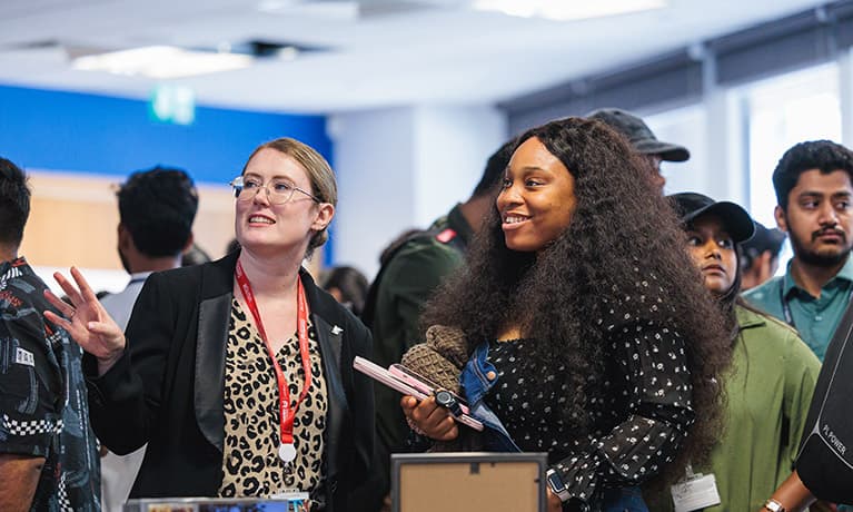 A lecturer talking to a student in a busy room of people