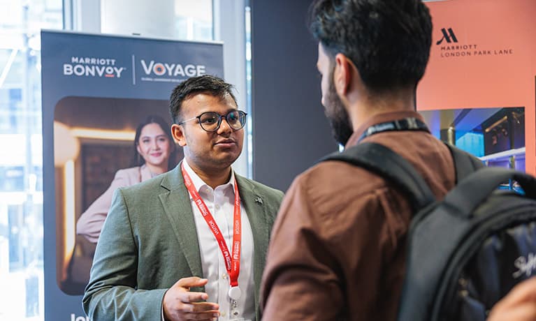 A male professional stood in front of a pull up banner talking to a student