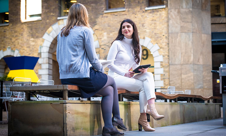 Two students sit chatting outside the London campus building.
