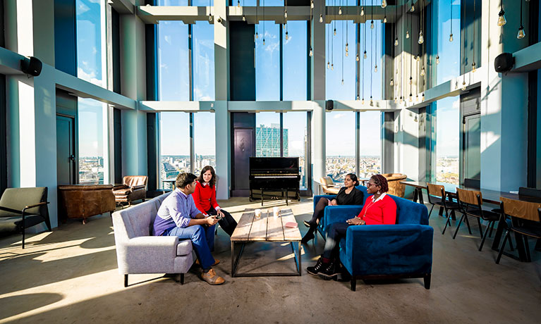 Students sitting in an open study area on campus, with tall windows behind them giving a skyline view over the city of London.
