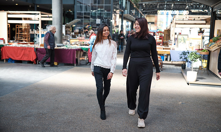 two females walking through an outdoor market with market stalls behind