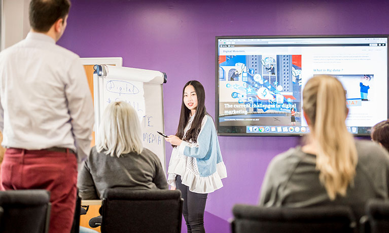 lady standing up presenting to three people  using a flip chart and presentation on screen
