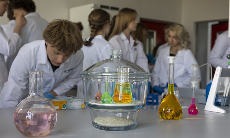 Young people in white lab coats next to a table with glasses of pots of different coloured liquids