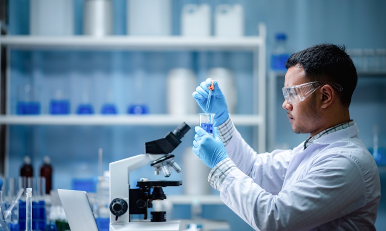 A researcher in a lab coat and protective goggles next to a microscope