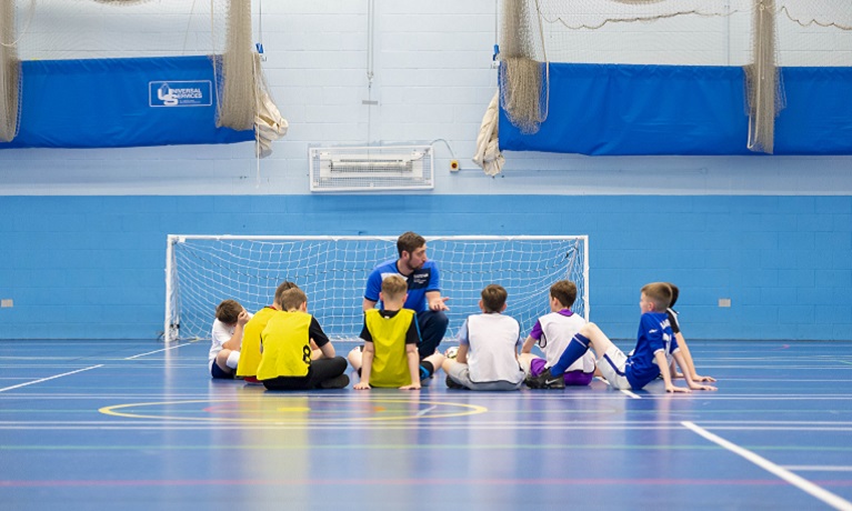 Young footballers sat listening to instructor in sports hall.