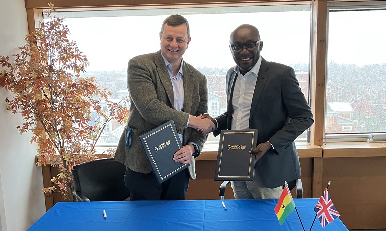 Richard Wells, Coventry University's Pro-Vice-Chancellor (International), and Dr Kingsley Agyemang, Registrar at Ghana Scholarship Secretariat, shaking hands while each holding a signed copy of the agreement and standing behind a table with a blue covering