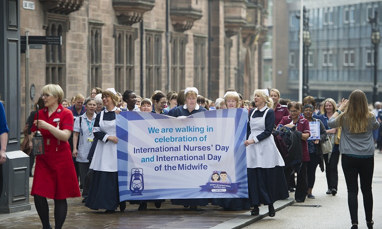 Group of ladies dressed like nurses walking in a city centre carrying a big banner that reads we are walking in celebration of International Nurses’ Day