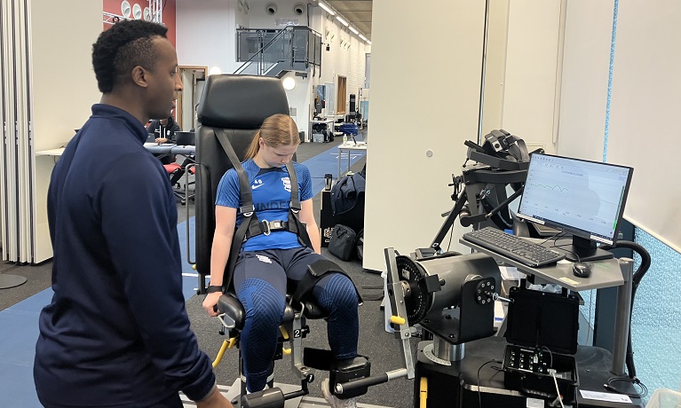 Birmingham City player Delphi Cole sat in a strength testing seat next to a computer showing the results of her testing while a student watches on