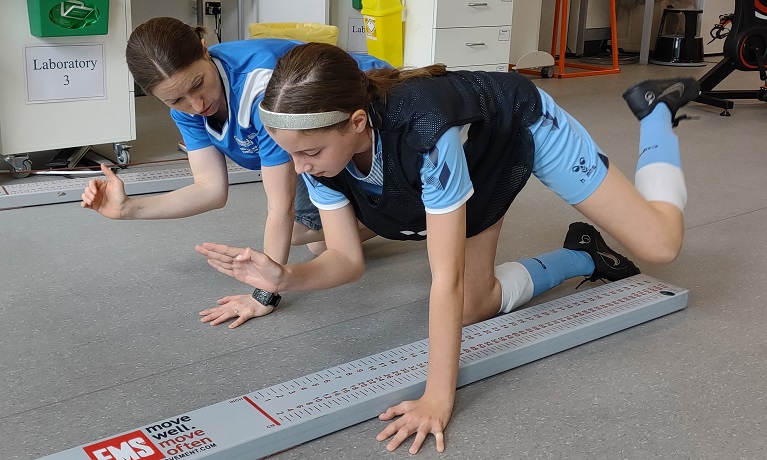 A girl wearing a football kit beside a beside a female sports science expert, being instructed how to do an exercise