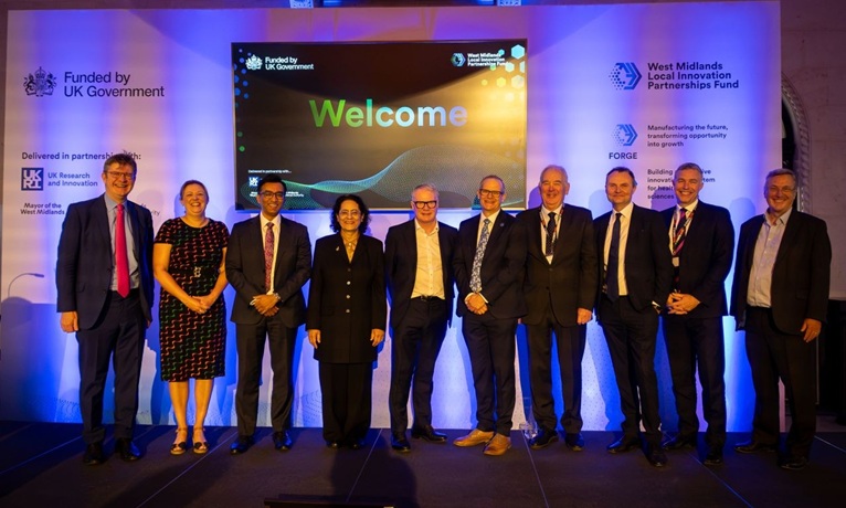 Ten people on a stage smiling at the camera with a backdrop for the West Midlands Local Innovation Partnerships Fund