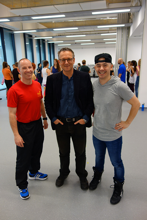 Three males stood in a busy room smiling