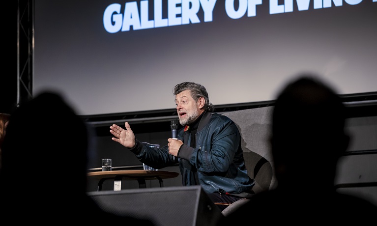 Actor Andy Serkis sitting on a chair on a stage speaking into a microphone