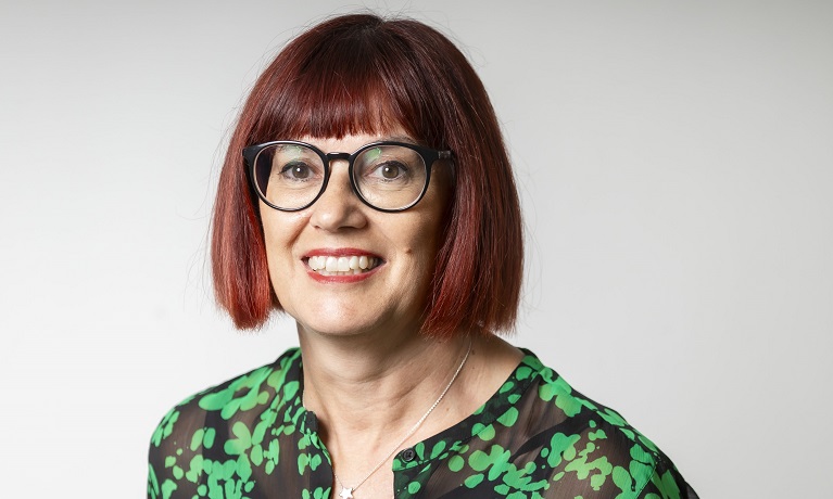 A head and shoulders image of Dr Chris Grant who has shoulder length reddy-brown hair and is wearing black glasses and a green and black dress