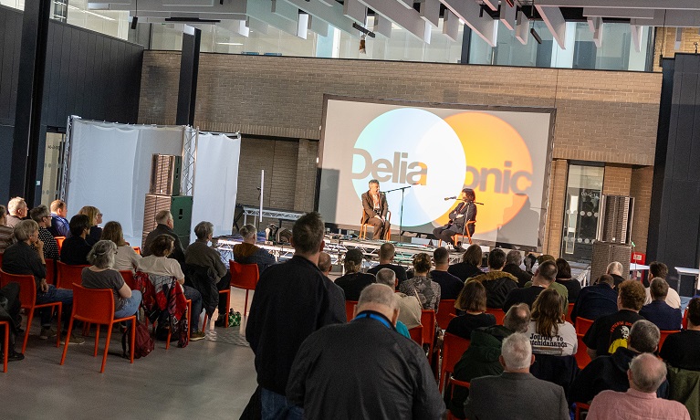 A crowd look on as Stuart Maconie and Cosey Fanni Tutti chat on a stage inside the Delia Derbyshire building