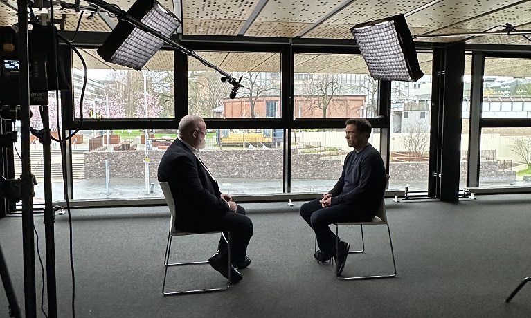 Two men sitting across from each other in from of camera equipment